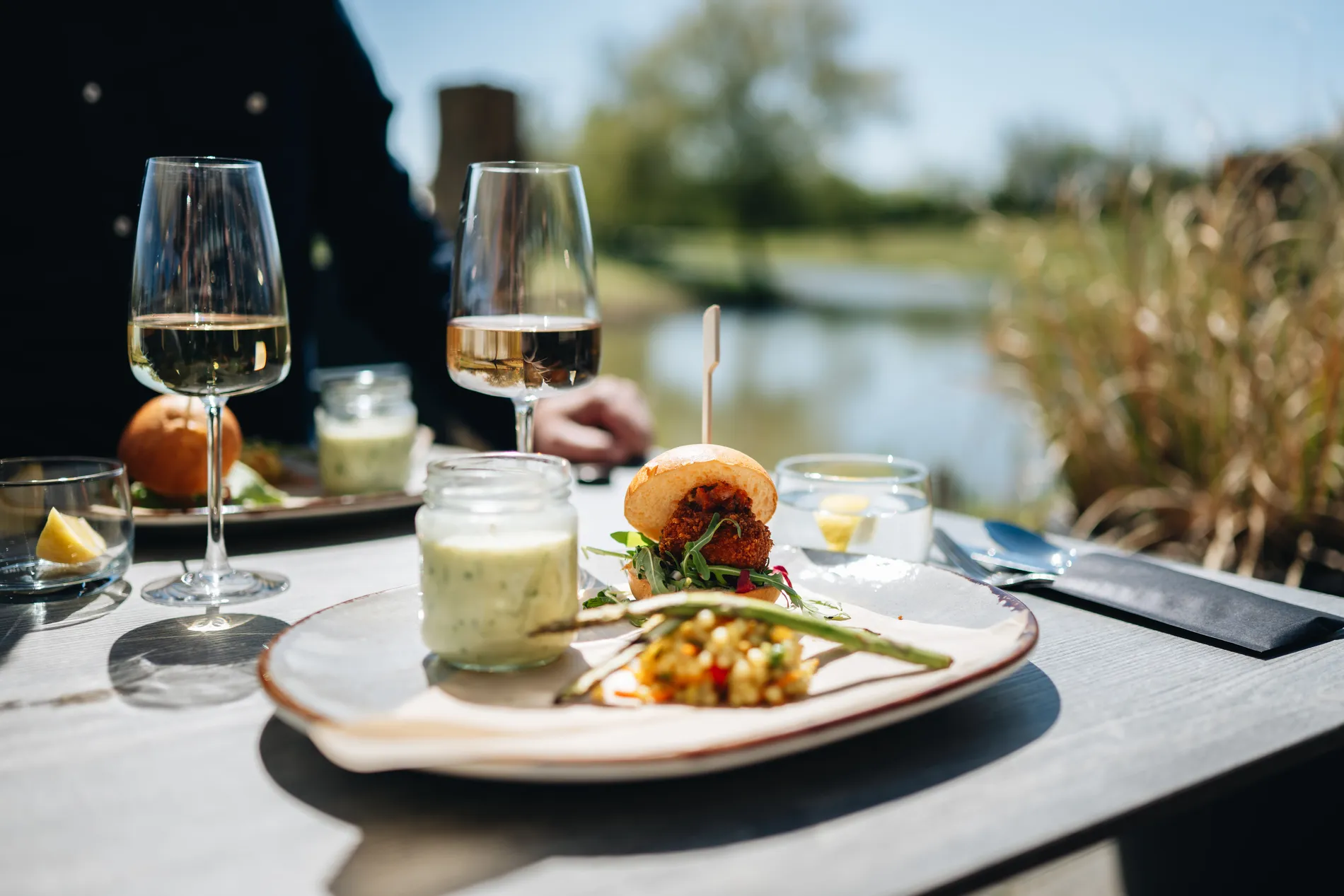Close-up van een lunchgerecht op een buitentafel met een mini-burger op een briochebroodje, een glazen potje met dressing en twee glazen witte wijn op de achtergrond aan het water.
